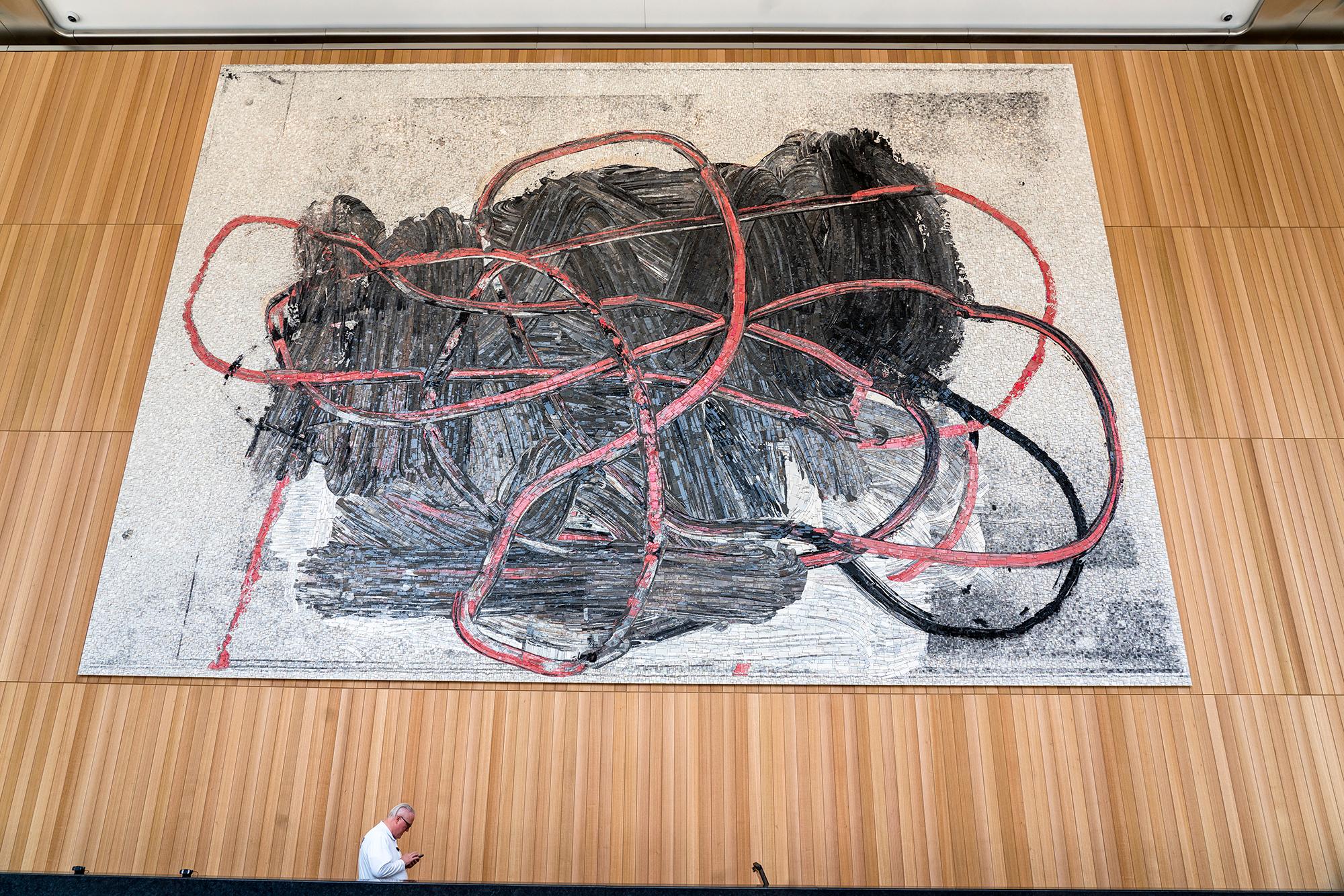 A color image of a mosaic installed in an interior space. The mosaic features an abstract arrangement: a salmon-colored curved line drawing laid over a black form on a white rectangle. The angle of the image is askew, capturing both the monumentality of the mosaic and for scale, a profile of a person cropped at the waist in the foreground.