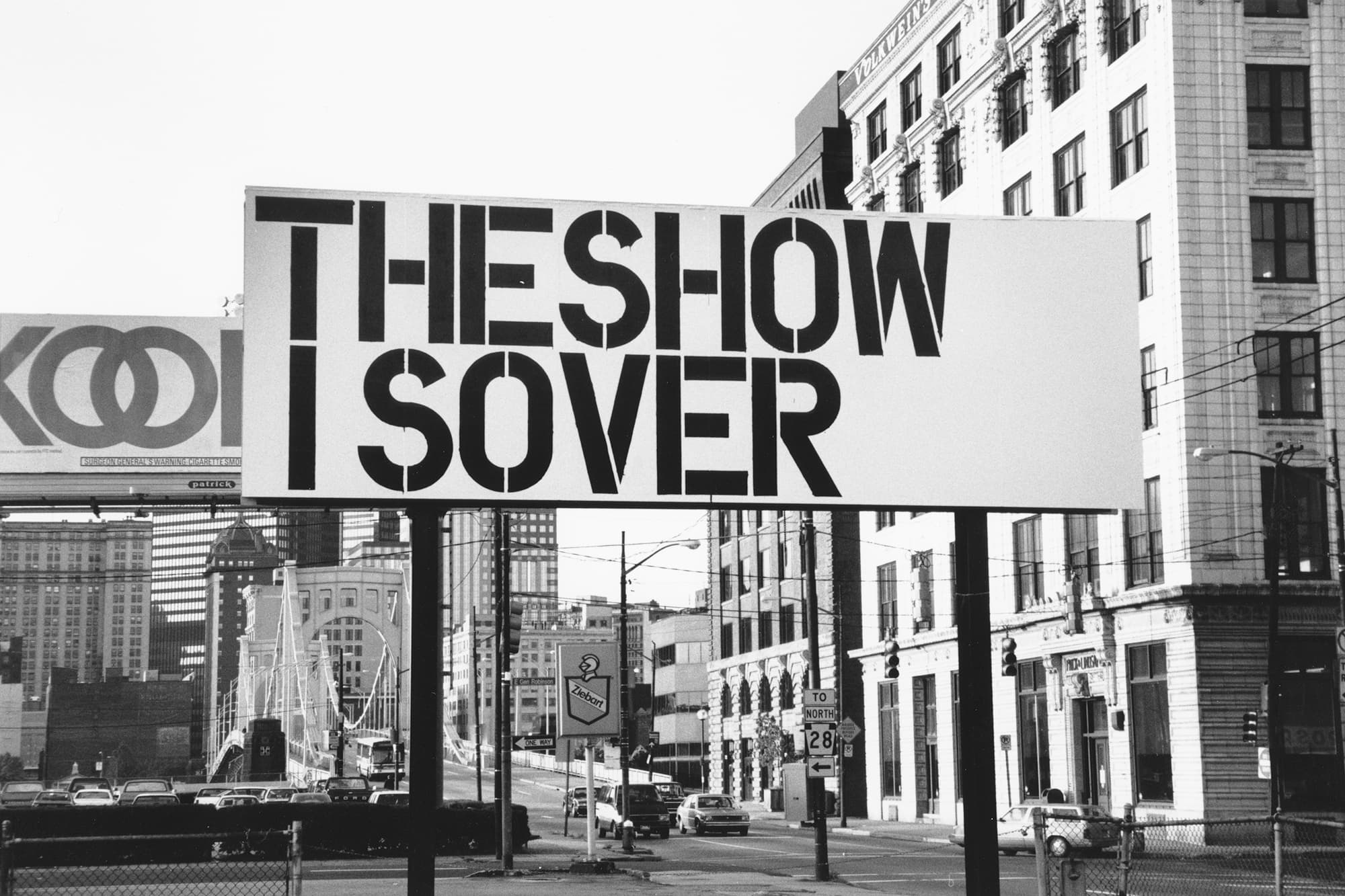 A black and white photograph shows a freestanding white billboard with black capitalized text: THESHOW atop the letters ISOVER. The billboard stands in an urban environment with a bridge and another billboard behind it with an ad for Kool cigarettes.