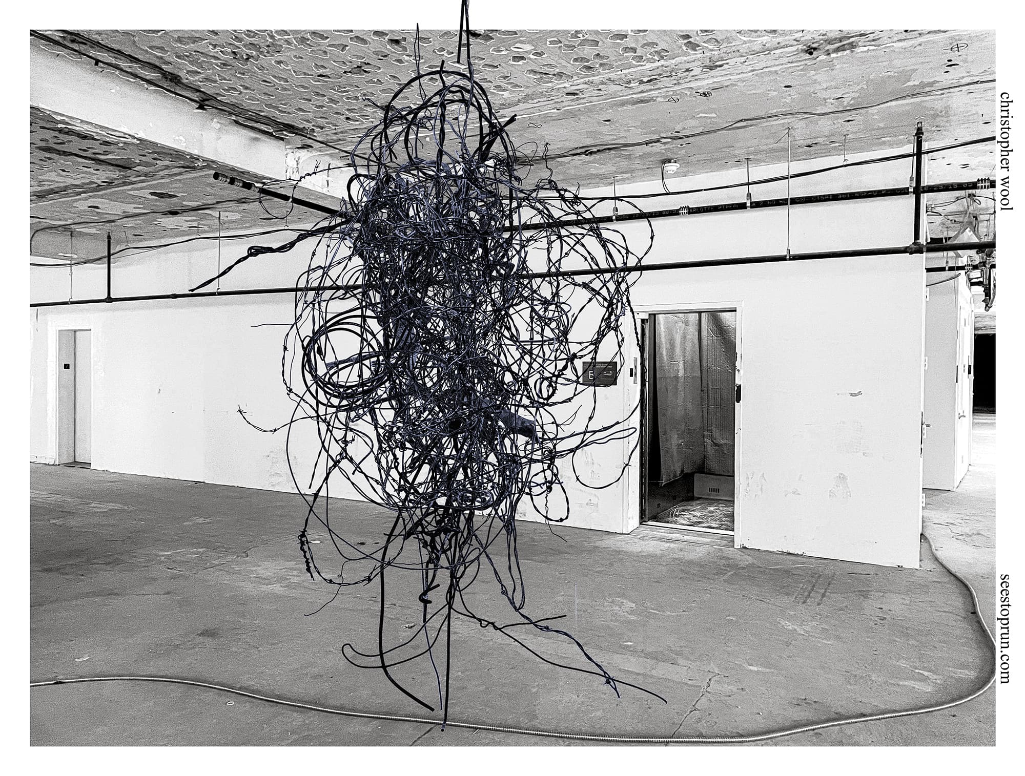 A two-tone image of a horizontally oriented exhibition poster. At center, a large tangled barbed wire sculpture, digitally colored in blue, hangs in front of an open elevator in an interior industrial space. The background is black and white. A long extension cord runs along the whole bottom of the image.