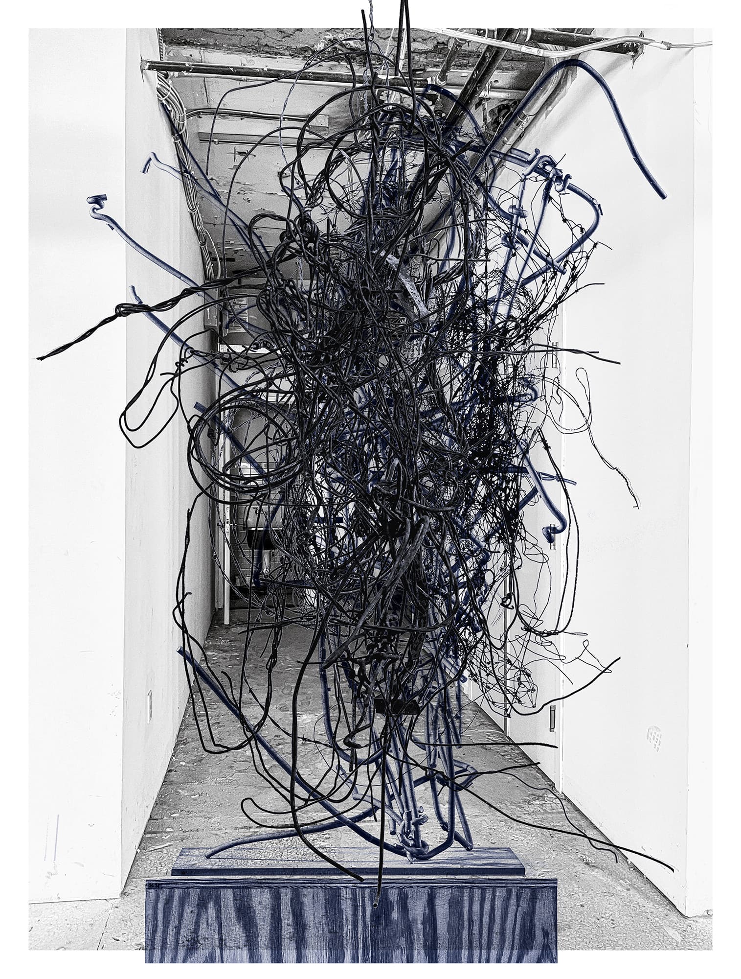 A two-tone image of a vertically oriented exhibition poster. A wire sculpture sits on a plywood pedestal in front of a hallway. The pedestal and sculpture are digitally colored blue and the background is black and white.