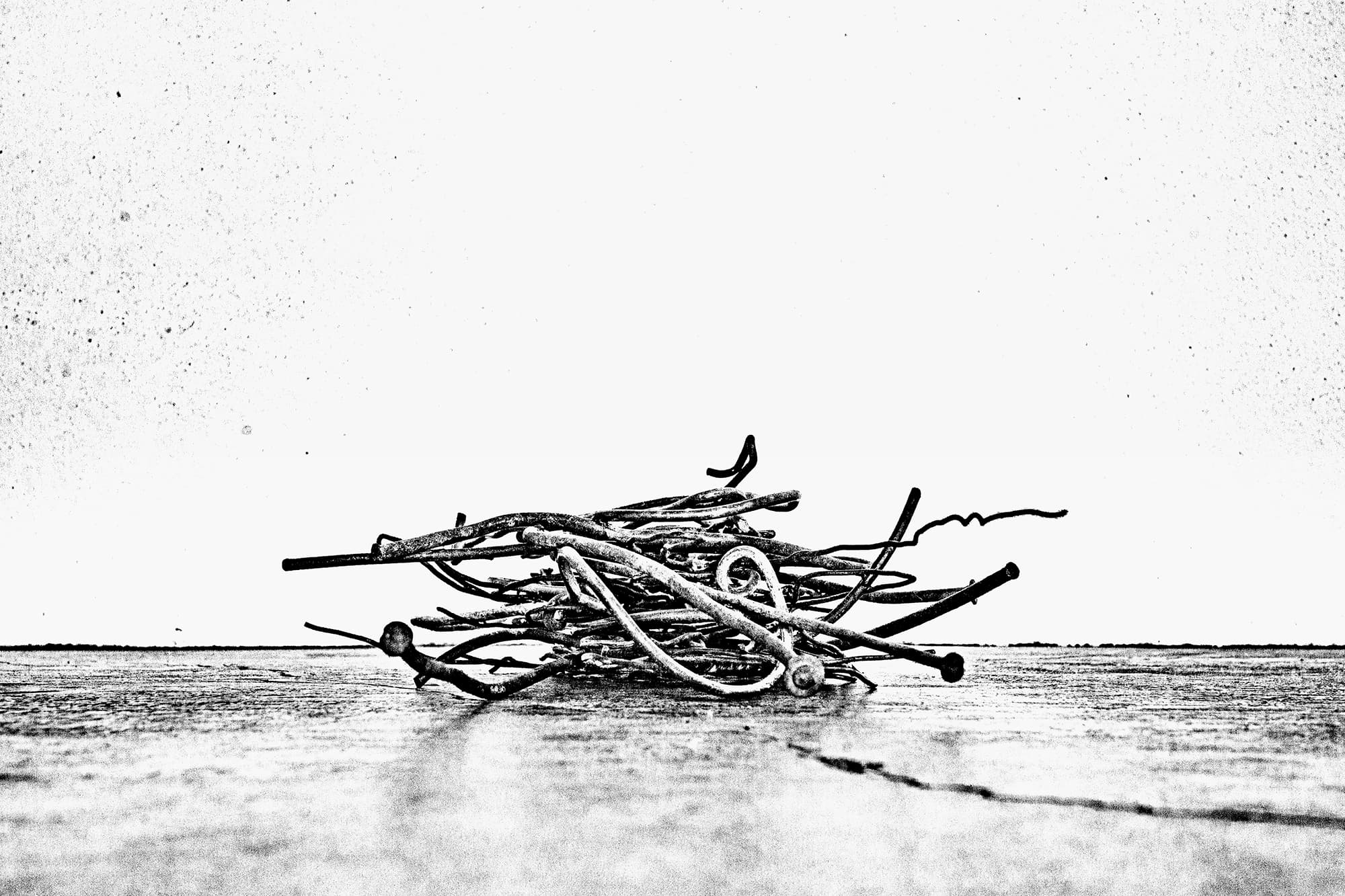 A black and white image of bent nails and wire on a floor-like surface. The grain of the photograph is distorted and a dappled effect is retained on all of the subjects in the image.