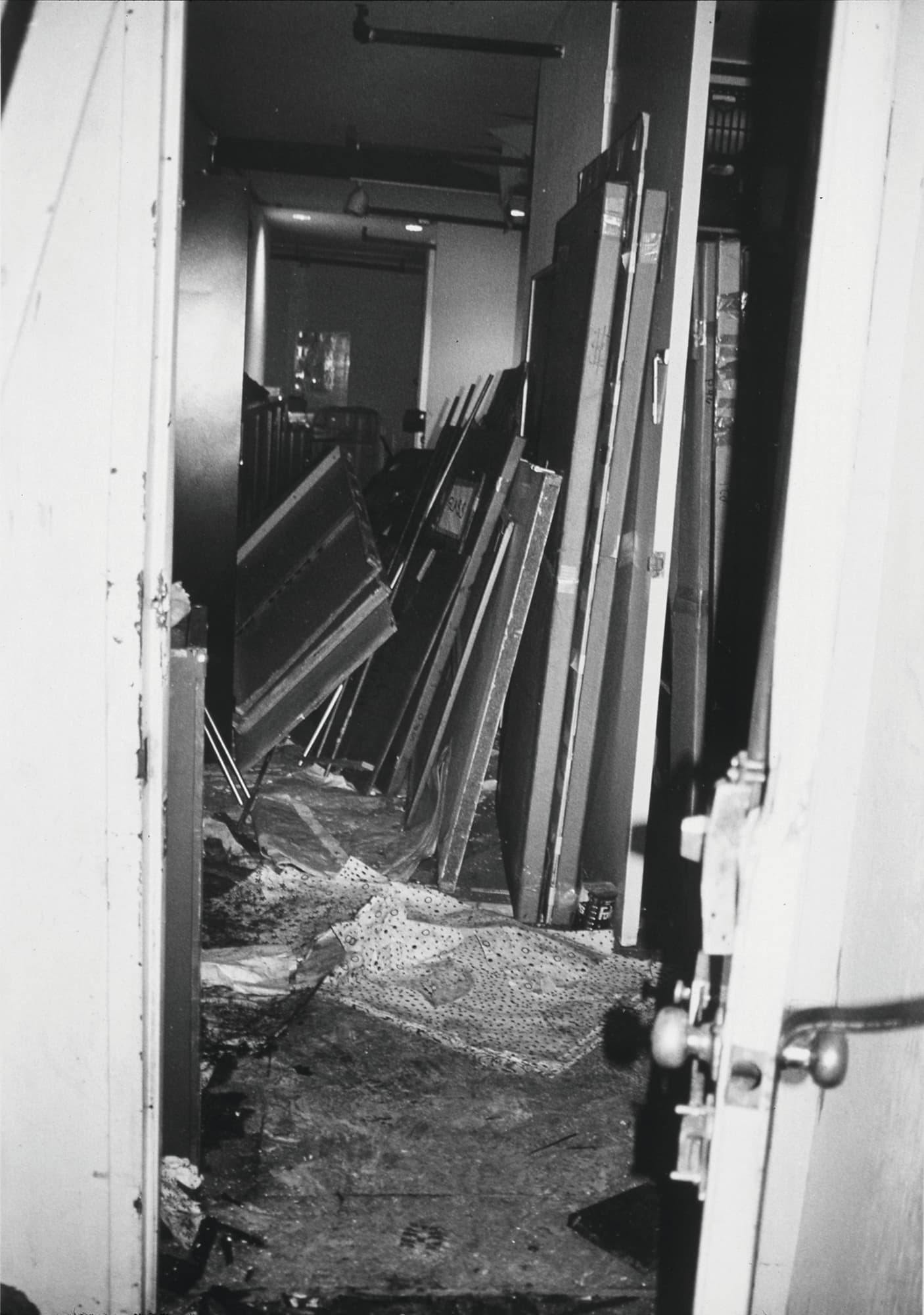 A black and white photograph. In the foreground, an open door washed out by a camera flash, is out of focus. Through the open door, in a dark corridor, large stacks of flat materials lean messily on interior walls.