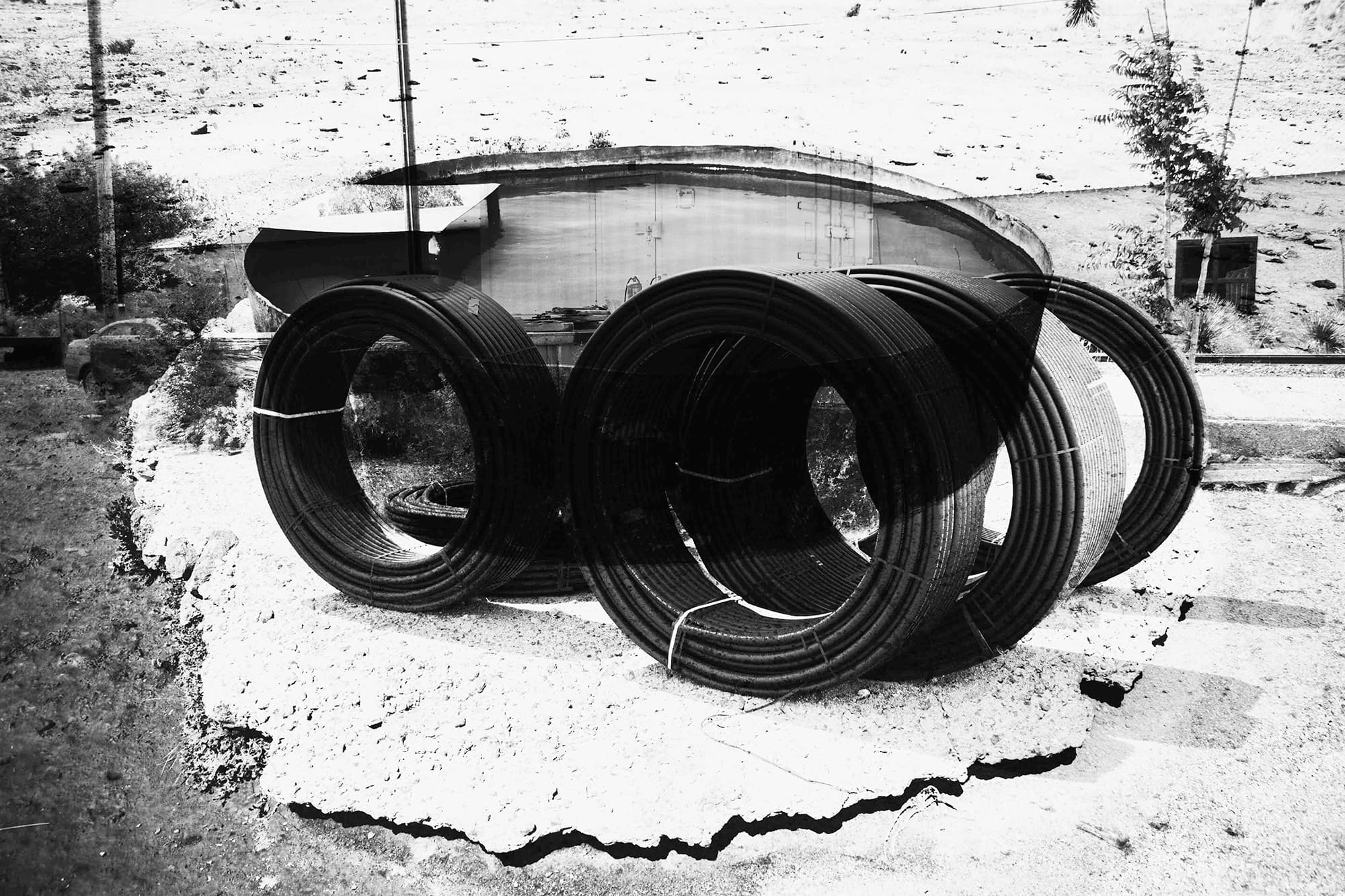 A black and white double exposure photograph features large curved shapes and dark circles. The circles appear to be made of large spools of bound pipe. A large singular abstract shape, that looks like an above ground pool, overlays the pipe forms. A torn shape arcs along bottom of the image.